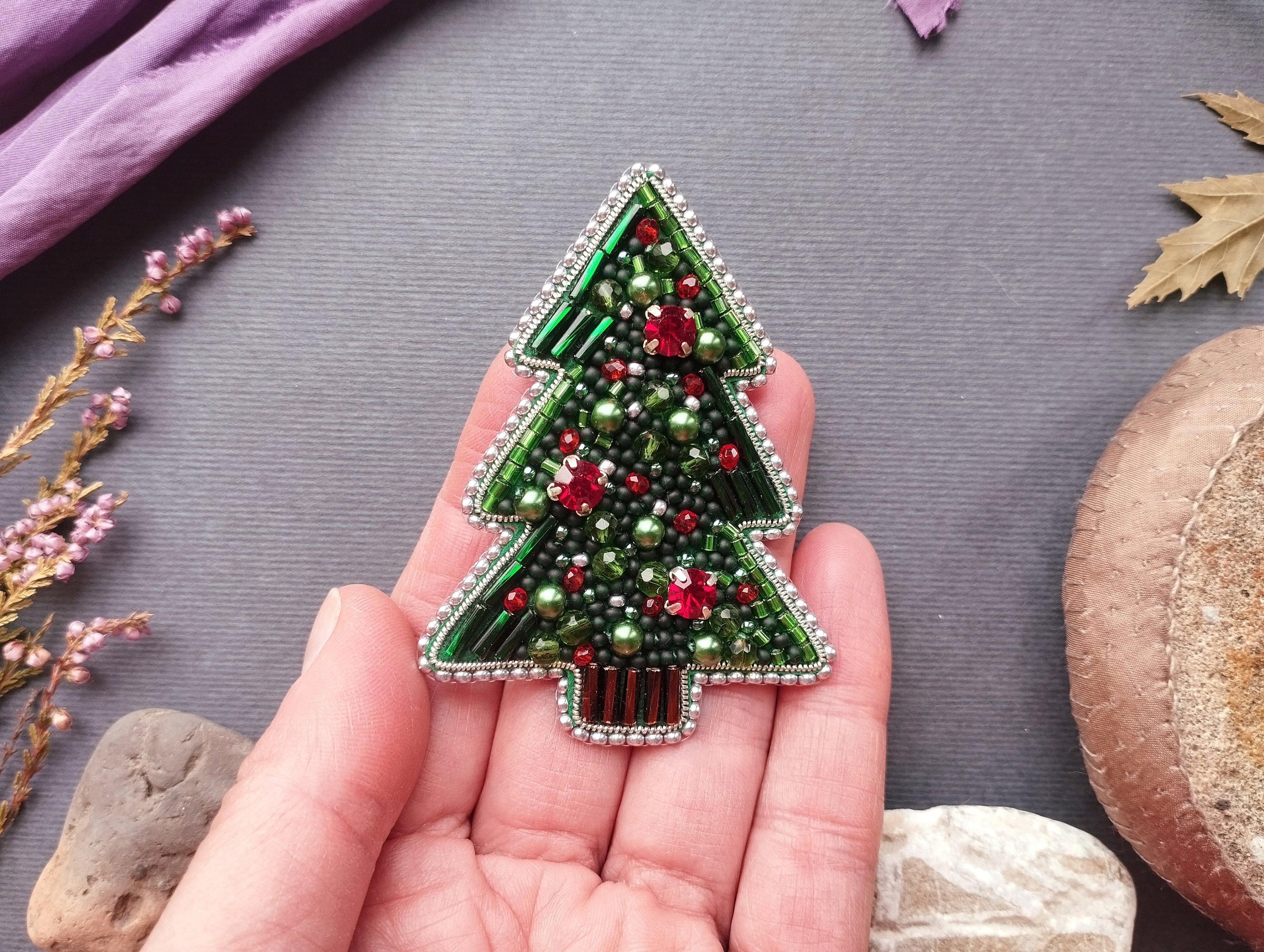 A hand holding a decorative Christmas tree brooch made of green beads and red and green beads, with a purple fabric and some leaves in the background.